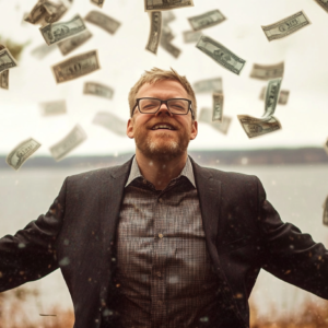 Man wearing glasses and a suit with dollar bills falling around him outdoors.