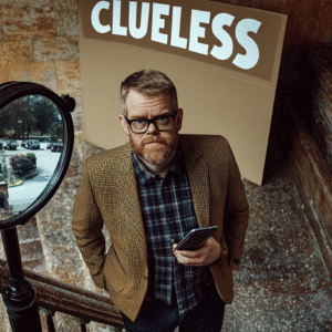 Man with glasses and a brown blazer holding a phone, standing near a mirror and a sign that says 'Clueless'.