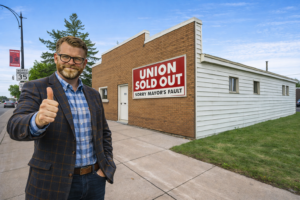 Portrait of Jim Paine-inspired figure wearing a blue plaid blazer and glasses standing beside a brick building with a bold UNIONS SOLD OUT sign on a sunny outdoor street scene