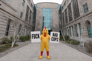 In a courthouse courtyard, a yellow mascot with red boots holds protest signs reading “FUCK” and “BAD COPS” amid civil rights storm clouds.