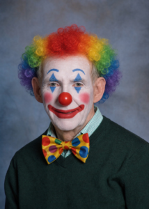 Jack Sweeney wears rainbow clown makeup, a red nose and a yellow polka dot bow tie against a muted studio backdrop.
