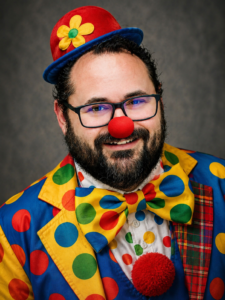 A smiling clown in a bright polka-dot suit, red nose and flowered hat wears glasses against a gray studio background.